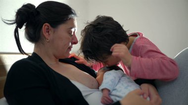 Büyük kardeş yeni doğmuş bebeği şefkatle öperken anne emzirirken sevgi, sıcaklık ve anne ile kardeş arasındaki bağ ile dolu dokunaklı bir aile anını yakalıyor.