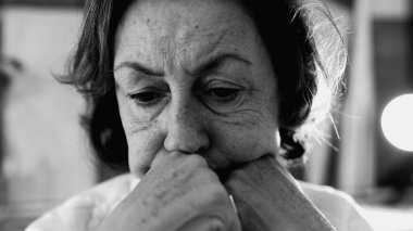 Kaşları çatık ve yüzünde derin çizgiler olan, yumruklarının üzerinde çenesi dinlenen, gözleri yere bakan yaşlı bir kadının yakın çekimi, siyah ve beyaz.