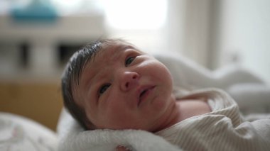 Yeni doğmuş bebek yukarı bakıyor, ağzı hafifçe açık, yumuşak bir battaniyeye sarılmış, annesinin kollarında rahat ve güvenli, parlak gözler, erken yaşamın mucizesini ve masumiyetini yakalıyor, huzurlu bir atmosfer. 