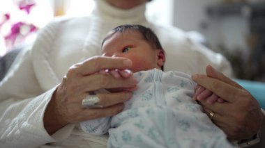 Yeni doğmuş bebek, büyük ebeveynlerinin elinde tutularak dünyaya merakla bakıyor. Farklı nesiller arasında paylaşılan değerli bağı ve sevgiyi vurguluyor.