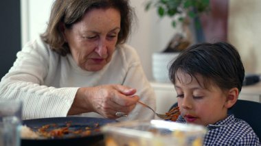 Büyükanne masada gönülsüz bir genci nazikçe besler, aile yemeği sırasında bir anlık ilgi ve bağlantı hisseder.