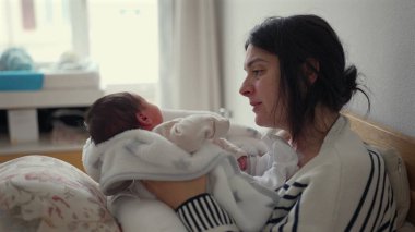 Genç anne yeni doğmuş bebeğine sevgiyle bakıyor, bebeği kollarına alıyor, hassas bir bağ anı, erken annelik, evde bağ kurma, yumuşak ışık, sıcak atmosfer.