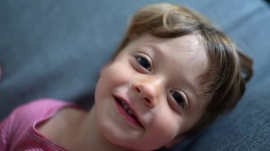 Kanepede uzanmış gülümseyen bir çocuk, neşe ve sıcaklıkla yukarı bakıyor. Rahat bir evde çocukluğun masumiyeti ve mutluluğu.