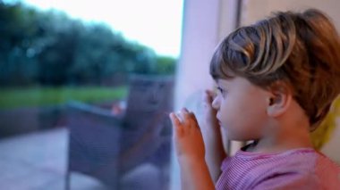 Elleri yüzün üzerinde, merakla ve heyecanla camdan dışarı bakan bir çocuk. çocukluğunda sessiz bir merak ve yansıma anı