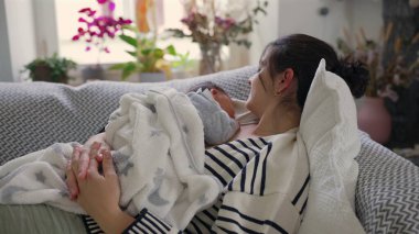 Anne yeni doğmuş bebeğini kucağında tutarken bir kanepede dinleniyor. Huzurlu bir ev ortamı ile çevrili. Aralarındaki rahatlık, güvenlik ve derin bağın altını çiziyor.