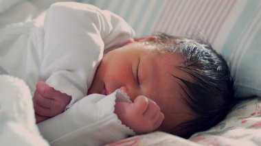 Yeni doğmuş bebek yatakta mışıl mışıl uyuyor, kolları içeri sokulmuş, sıcaklıkla dolu sıcak ve güvenli bir ortamda ilk yaşamın şefkatini ve sükunetini gösteriyor.