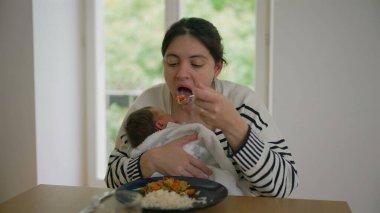 Anne, yeni doğmuş bebeğini yanında tutarak öğle yemeği yiyerek, parlak bir pencerenin kenarındaki masaya oturarak, zorlukları ve anneliği günlük rutinlerle dengeleyerek,