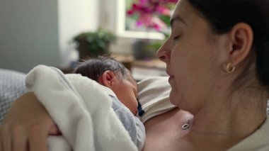 Anne yeni doğmuş bebeğini kucaklarken esniyor, erken ebeveynliğin yorgunluğunu ve hassas anlarını gösteriyor, zorluklara rağmen paylaşılan bağ ve sevgiyi vurguluyor