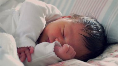 Yeni doğmuş bebek yatakta mışıl mışıl uyuyor, kolları içeri sokulmuş, sıcaklıkla dolu sıcak ve güvenli bir ortamda ilk yaşamın şefkatini ve sükunetini gösteriyor.