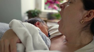 Anne yeni doğmuş bebeğini kucaklarken esniyor, erken ebeveynliğin yorgunluğunu ve hassas anlarını gösteriyor, zorluklara rağmen paylaşılan bağ ve sevgiyi vurguluyor