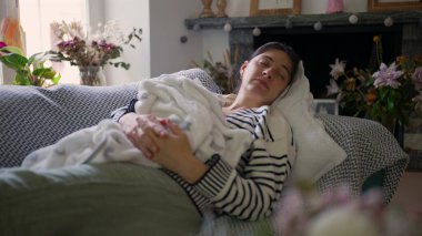 Anne bir kanepeye uzanıyor, gözleri kapalı, yeni doğmuş bebeğini bir battaniyeye sarmış, sakin, rahat bir atmosferle çevrili, odada süzülen yumuşak doğal ışıkla.