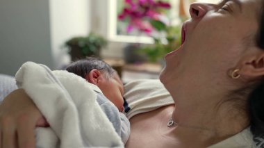 Anne yeni doğmuş bebeğini kucaklarken esniyor, erken ebeveynliğin yorgunluğunu ve hassas anlarını gösteriyor, zorluklara rağmen paylaşılan bağ ve sevgiyi vurguluyor