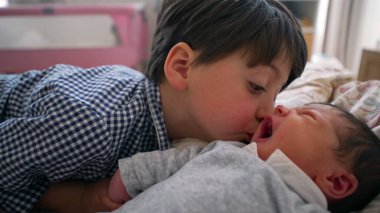 Küçük çocuk, yeni doğan kardeşinin elini tutarken yatakta sıcak bir şekilde gülümsüyor. Sevgi dolu ve şefkatli bir aile ortamında kardeş bağı kurmanın masumiyetini ve saf neşesini yakalıyor.