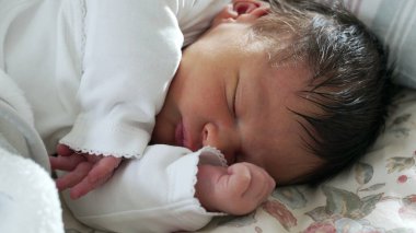 Koyu renk saçlı yeni doğmuş bebek çiçek yatağında mışıl mışıl uyuyor, kolları içeri sokulmuş, sıcaklıkla dolu sıcak ve güvenli bir ortamda ilk yaşamın şefkatini ve huzurunu sergiliyor.