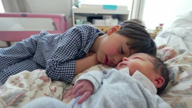 Küçük bir çocuk, bir yatakta uzanırken yeni doğan kardeşini yanağından öpüyor. Kardeşler arasında gelişen sevgi ve bağın yansıttığı şefkatli bir anı yakalıyor.