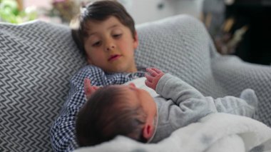 Büyük kardeş, sıcak bir aile ortamında derin bir kardeşlik bağı kurmaya başladıklarında, onları nazikçe tutarken, bir anlık merak ve yakınlık göstererek, yeni doğan kardeşe dikkatle bakıyor.
