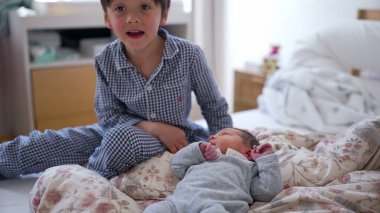 Genç çocuk gülümsüyor ve yeni doğan kardeşinin yanında oturuyor. Yeni bir aile üyesinin mutluluğunu ve heyecanını yansıtan neşeli ve iç açıcı bir sahne yaratıyor.