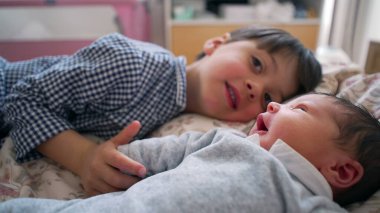 Küçük çocuk, yeni doğan kardeşinin elini tutarken yatakta sıcak bir şekilde gülümsüyor. Sevgi dolu ve şefkatli bir aile ortamında kardeş bağı kurmanın masumiyetini ve saf neşesini yakalıyor.