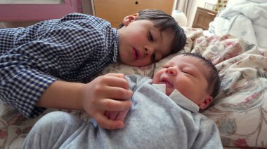 Küçük çocuk yeni doğan kardeşinin yanında yatıyor, neşe içinde gülümsüyor, yakın bir aile ortamında kardeşler arasındaki saf mutluluk ve sevgiyi vurguluyor.