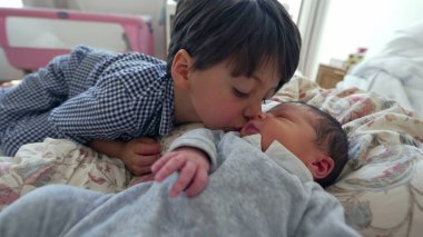 Küçük bir çocuk, bir yatakta uzanırken yeni doğan kardeşini yanağından öpüyor. Kardeşler arasında gelişen sevgi ve bağın yansıttığı şefkatli bir anı yakalıyor.