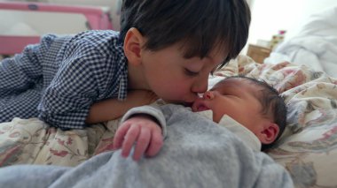 Küçük bir çocuk, bir yatakta uzanırken yeni doğan kardeşini yanağından öpüyor. Kardeşler arasında gelişen sevgi ve bağın yansıttığı şefkatli bir anı yakalıyor.