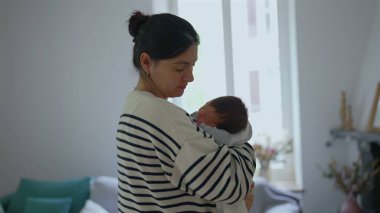 Sessiz bir odada, yeni doğmuş bebeğini nazikçe kucaklayan bir annenin, anne-baba ve çocuk arasındaki samimiyet, güvenlik ve derin bağın altını çizdiği görüntüsü.