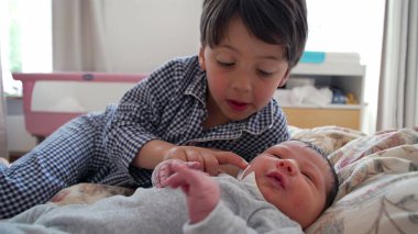 Ağabeyi, yeni doğan kardeşiyle yatakta etkileşim halinde. Sıcak, aile odaklı bir evde kardeşler arasındaki özel bağ ve ilgiyi vurguluyor.
