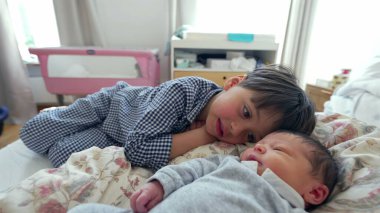 Küçük bir çocuk, bir yatakta uzanırken yeni doğan kardeşini yanağından öpüyor. Kardeşler arasında gelişen sevgi ve bağın yansıttığı şefkatli bir anı yakalıyor.