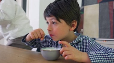 Yemek masasında yoğurdun tadını çıkaran genç bir çocuk evde sakin bir yemek yerken, çocukluğun basit zevklerini vurguluyor.