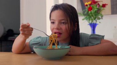 Çatalla makarna yiyen genç bir kız, yemeğine odaklanmış, aile yemeklerinde evde sakin bir anın tadını çıkarıyor.