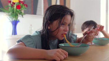 Masada odaklanmış bir ifadeyle makarna yiyen genç bir kız, arka planda yemek yiyen bir kardeş, her ikisi de evde rahat bir aile yemeğinin tadını çıkarıyor.