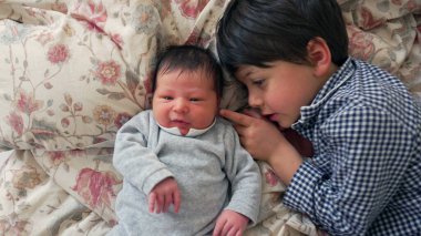 Ağabeyi çiçeklerle kaplı bir yatakta yeni doğan kardeşinin yanında sarılıyor. Huzurlu ve rahat bir yuvada kardeşçe sevgi ve bağlılık sahnesi yaratıyor.