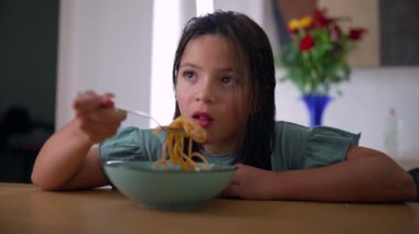 Genç bir kız kaseden makarna yiyor, bir ısırık alırken odaklanıyor, sakin ve rahat bir ortamda evde rahatça yemek yiyor.