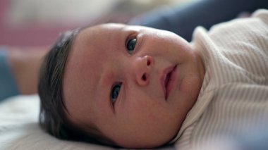 Geniş, meraklı gözleri ve yumuşak ifadeleri olan bir bebeğin yüzüne yakın çekim. Bebek sıcak bir ev ortamında bir yatakta yatıyor.