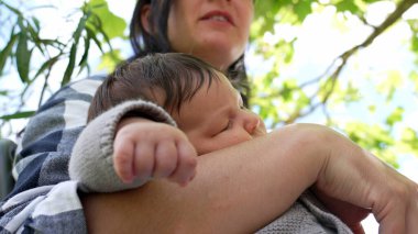 Annesinin kollarında dinlenen uyuyan bir bebeğin yakın plan çekimi. Bu arada anne, derin bir dokunuşla dudaklarına dokunuyor ve dışarıda sakin bir anın tadını çıkarıyor.