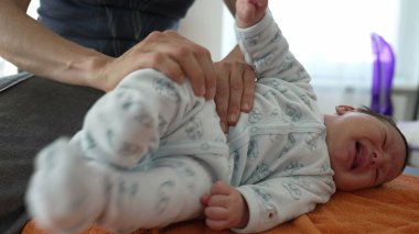 Ortopedist, yeni doğmuş bebeklerin bacaklarını turuncu bir havluyla nazikçe esneterek vücut ağrılarını dindiriyor. Bebek ağlamaya başlar, tedavi sırasında rahatsız hisseder..