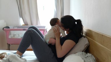 Anne bebeğini kucağında tutarken yatakta uzanıyor arka plandaki pencereden gün ışığıyla şefkatli bir öpücük paylaşıyor. Yumuşak ışık perdeleri ve pembe bebek beşiği olan rahat bir ev ortamı