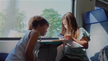 Trende birlikte oturan iki çocuk, kitap okuyup yazmaya odaklanmış, manzaralı bir gezide birlikte kitap okuyorlar.