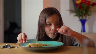 Yemek masasında yemek yiyen genç bir kız, ev ortamında tabağındaki yemekle ilgilenmek için çatal ve bıçak kullanmaya odaklanıyor.