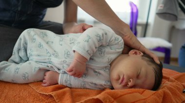 Ortopedist, çocukluğunda vücut ağrılarını hafifletmek için turuncu havluyla yeni doğan bebeğin pozisyonunu ayarlıyor, bebek rahat bir tulum giyiyor, dikkatli ve profesyonel yaklaşımı gösteriyor.