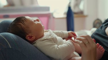 Yeni doğmuş bir bebeğin yüzüne yakın çekim, annesinin bacağından tutularak, bebeğin masumiyetini ve sükunetini yakalıyor, yumuşak ve rahat bir çevreyle çevrili.