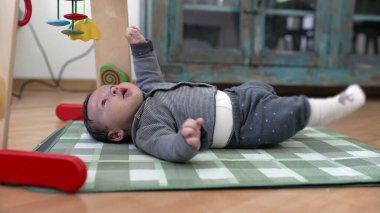 Kareli bir paspasın üzerinde uzanmış bebek tuhaf bir ifadeyle renkli oyuncaklara bakıyor. Sıcak bir ev ortamında neşeli bir doğa.