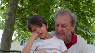 Elderly man and young boy focused on reading a book together outdoors, sharing a tender and educational moment under the shade of trees. intergenerational bonding clipart