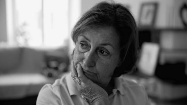 Düşünceli bir ifadeye sahip yaşlı bir kadın, elini çenesinde dinlendiriyor, ev ortamında, yumuşak ışıklandırmada, siyah ve beyaz bir anı yakalıyor.