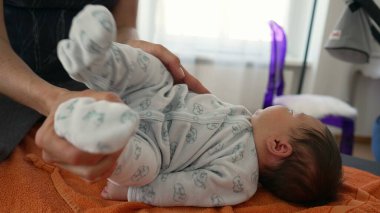 Ortopedist, çocukluğunda vücut ağrılarını hafifletmek için turuncu havluyla yeni doğan bebeğin pozisyonunu ayarlıyor, bebek rahat bir tulum giyiyor, dikkatli ve profesyonel yaklaşımı gösteriyor.