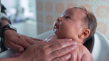 Bir küvette bebek, bakıcının elinden su damlarken dikkatle yukarı bakıyor, sıcak ve sıcak bir ortamda banyo sırasında bağ kurmanın hassas bir anını vurguluyor.