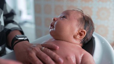 Bir küvette bebek, bakıcının elinden su damlarken dikkatle yukarı bakıyor, sıcak ve sıcak bir ortamda banyo sırasında bağ kurmanın hassas bir anını vurguluyor.