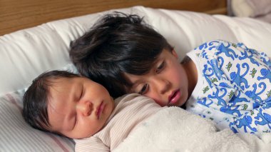 İki kardeşin birlikte uyuduğu yakın plan. Büyük olan, genç olanı nazikçe tutuyor. İkisi de yumuşak bir yatakta huzur içinde dinleniyor ve iç açıcı bir aile anı yaratıyor.