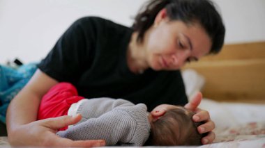Anne bebeğiyle bir yatakta dinleniyor, bebeği nazikçe kucaklıyor ve rahatlatıyor, huzurlu ve rahat bir ev ortamında sessiz ve sevgi dolu bir anı canlandırıyor.