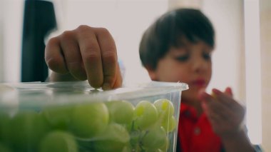 Genç çocuk dikkatle yeşil üzümlerden oluşan bir kabın içindeki plastik örtüyü soyuyor, odaklandığını ve evdeki basit, günlük aktivitelere odaklandığını vurguluyor.